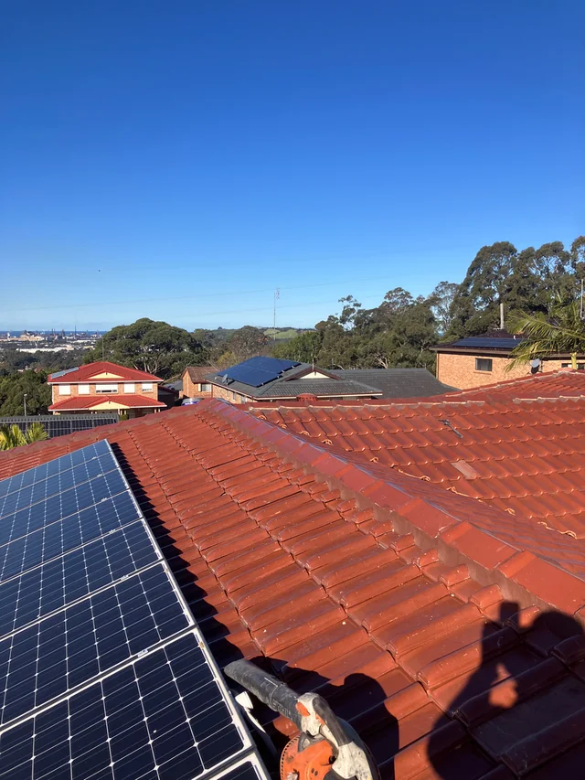 Red tiled roof beside solar panels, with neighbouring roofs and trees in the distance.